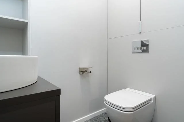 Moderní bílý záchod nainstalovaný v minimalistické koupelně s tmavým umyvadlem.