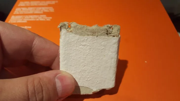 Rozbitý kus bílého azbestocementu v ruce na oranžovém pozadí.