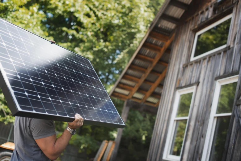 Muž nese fotovoltaický panel u starého dřevěného domu.