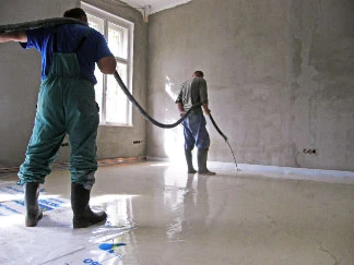 Dva dělníci nalévají tekutý betonový potěr do interiéru.