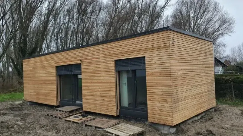 Moderní dřevěný modulový dům s velkými okny na nezpevněném pozemku.
