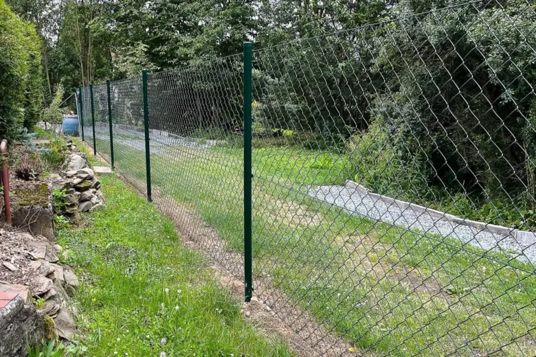 Zelená plot z pletivového drátu s kovovými sloupky v zahradě.