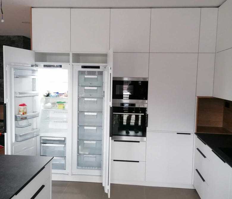 Bílá vestavná americká lednice otevřená v kuchyni s bílými skříněmi.