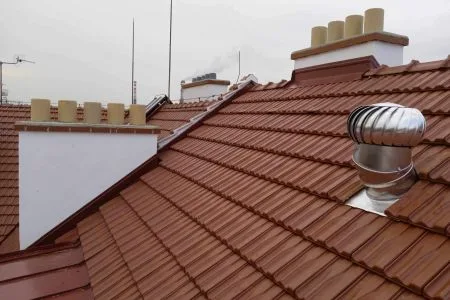 Kovový větrací komín na střeše s červenými taškami.