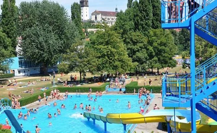 Venkovní bazén v Praze s kluzáky a lidmi koupející se na slunci.