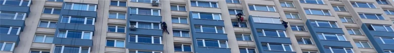 Pracovníci umývají fasádu šedého panelového domu.