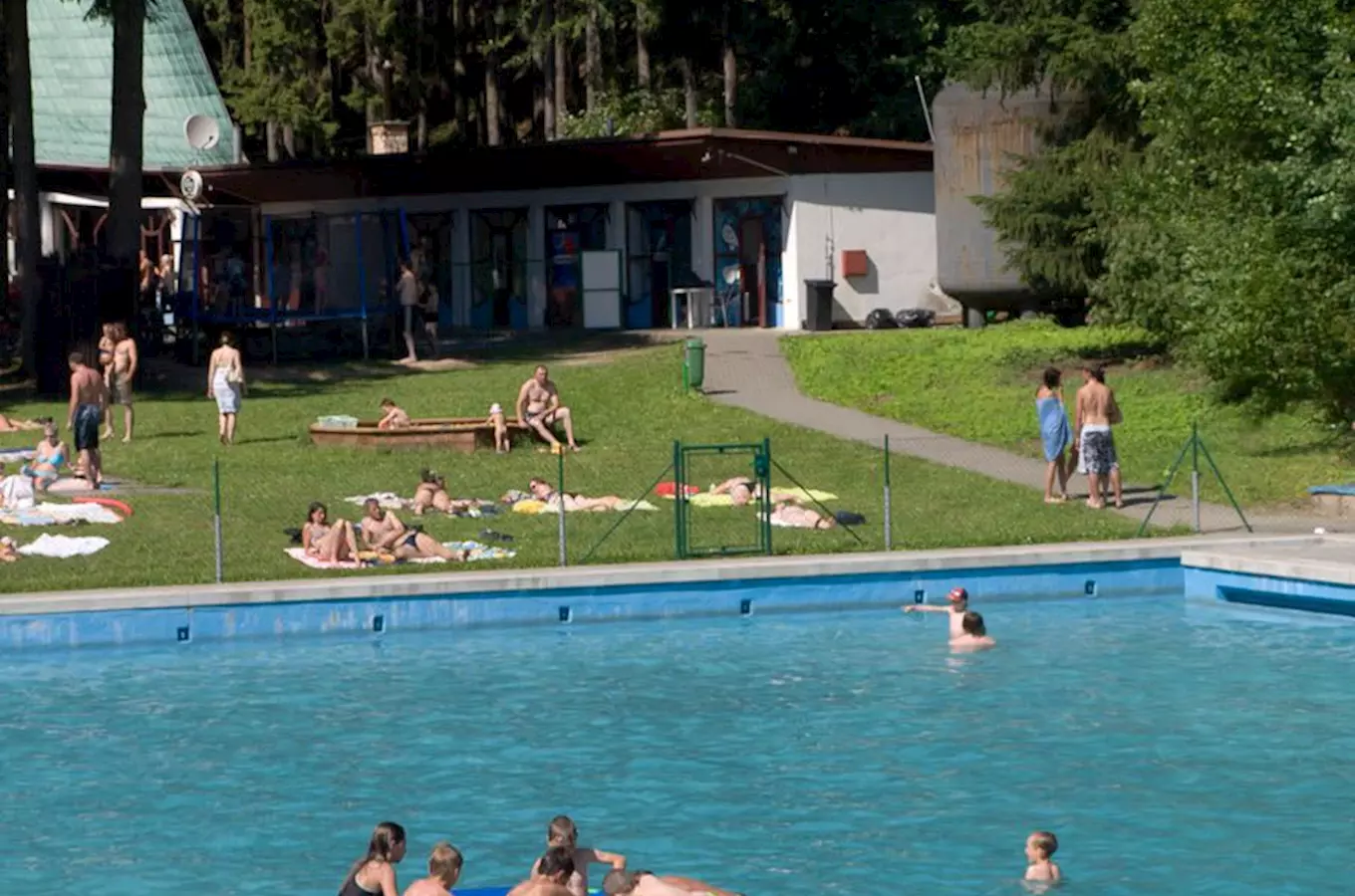 Venkovní bazén s lidmi, kteří se koupou a sluní na trávníku.