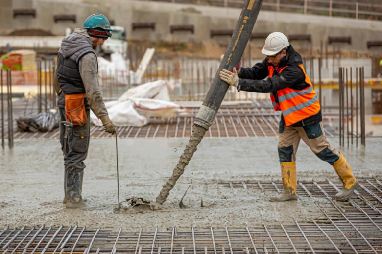 Dva dělníci nalévají beton přes výztužovou mřížku na stavbě.
