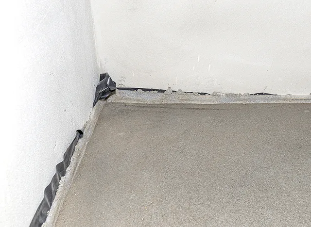 Detail dilatační spáry v rohu podlahy s černou páskou.