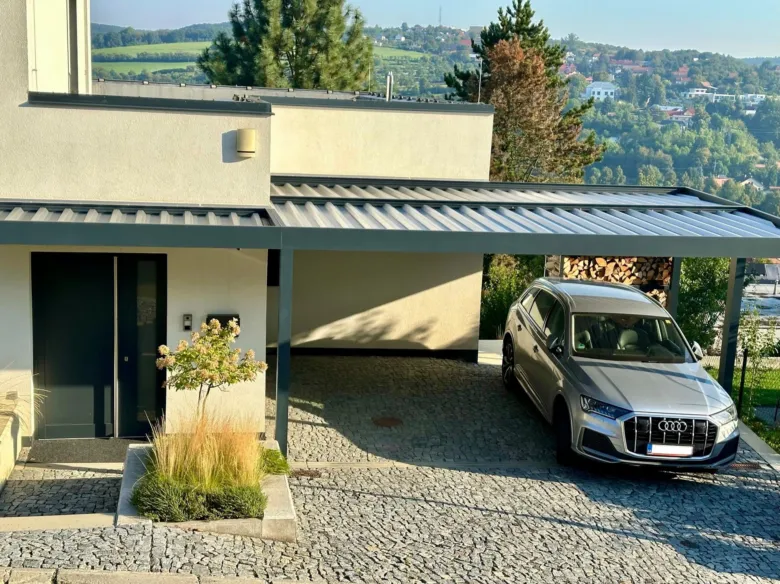 Stříbrný automobil pod kovovou pergolou u domu.