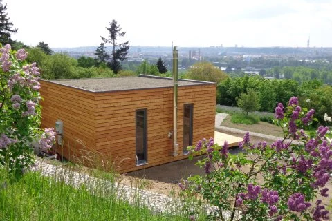 Moderní dřevěná modulární stavba na svahu s kvetoucími keři v popředí.