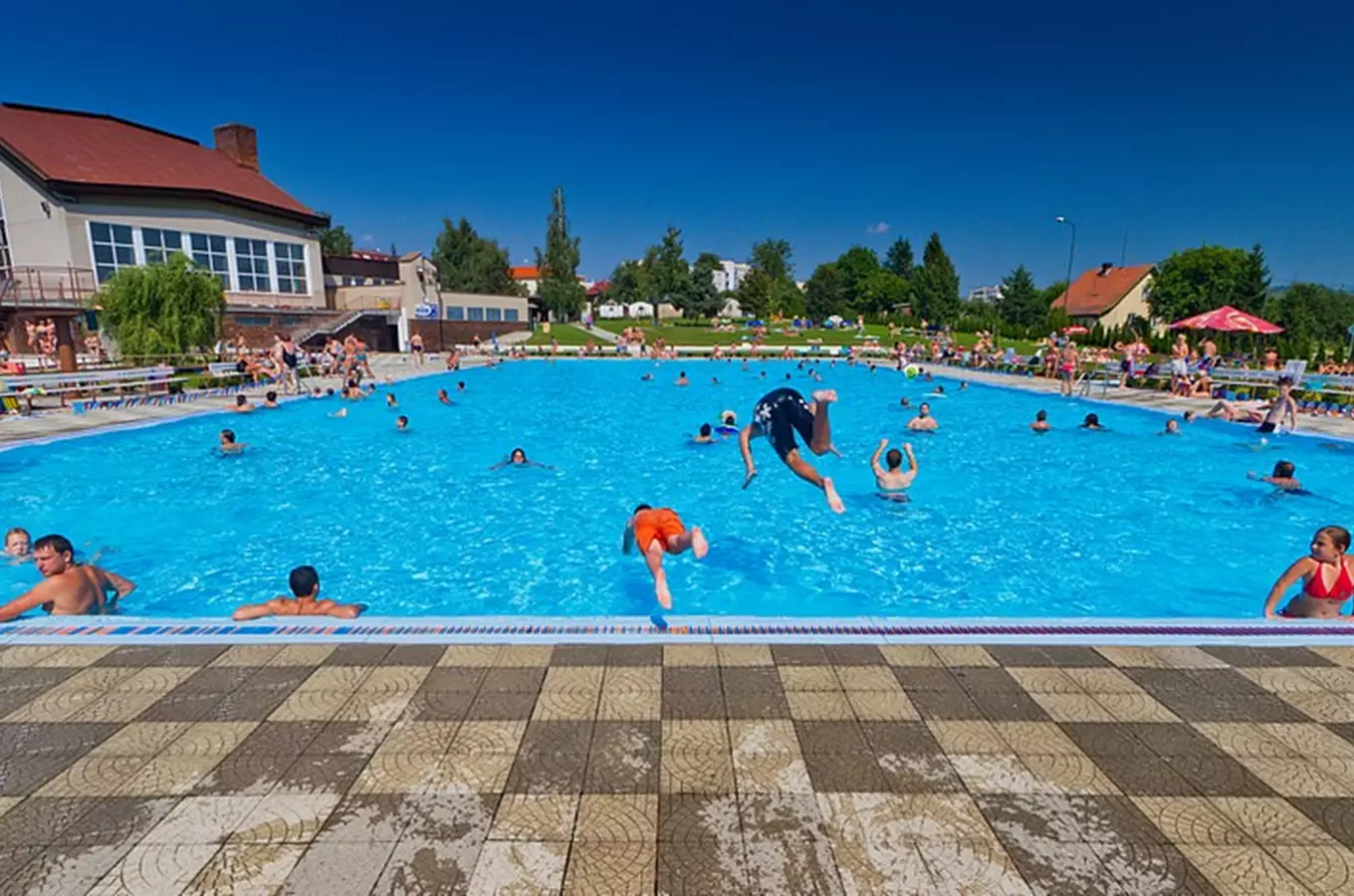 Lidé plavou v bazénu na slunečním dni s diváky kolem.