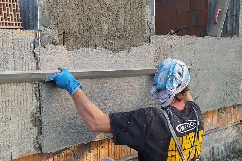 Stavebník hladí omítku na zdi pravítkem.