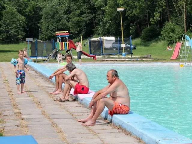 Lidé u venkovního bazénu s dětským hřištěm v pozadí.
