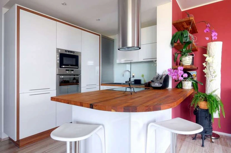 Moderní kuchyňský barevný pult s dřevěnou deskou, bílými židličky a kvetoucími orchidejemi na červené stěně.