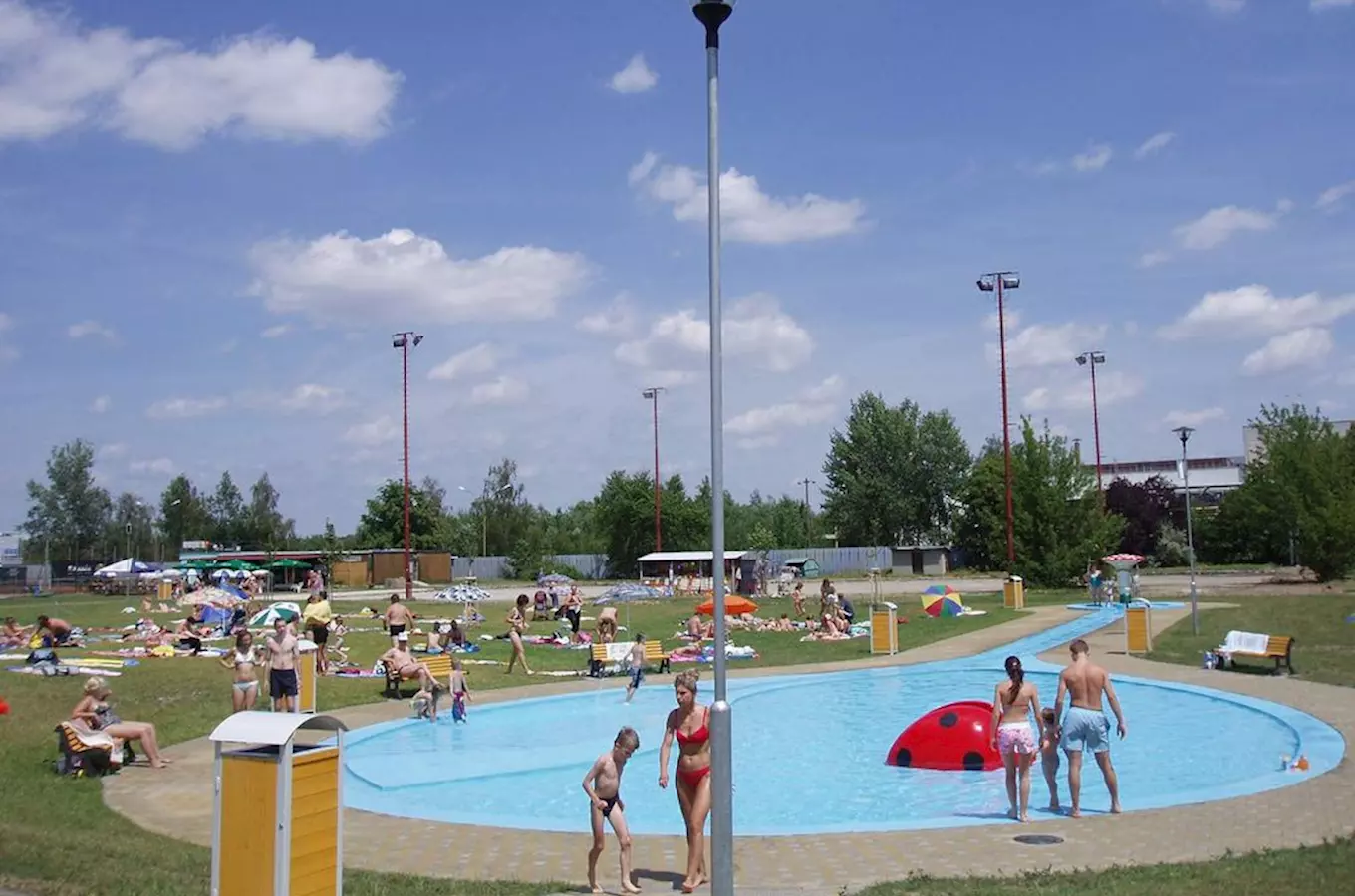 Venkovní bazén v koupališti Chropyně s lidmi plavajícími na slunci pod modrou oblohou.