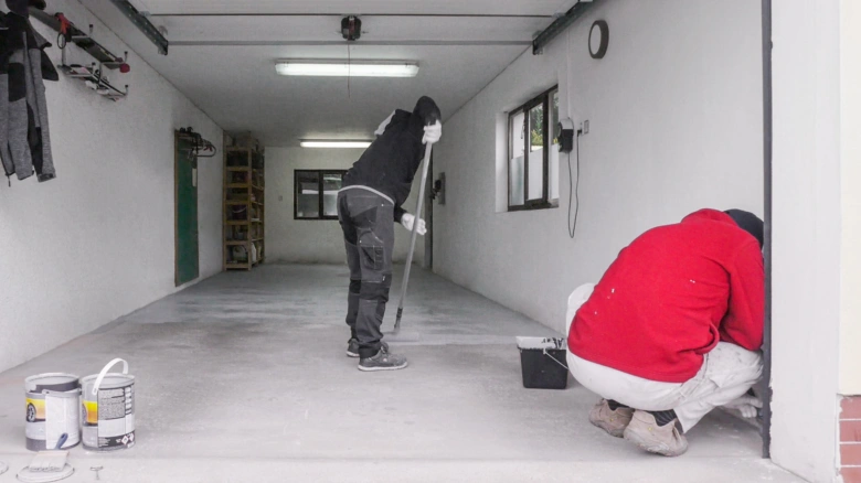 Dva lidé malují betonovou podlahu v garáži válečky a štětcem.