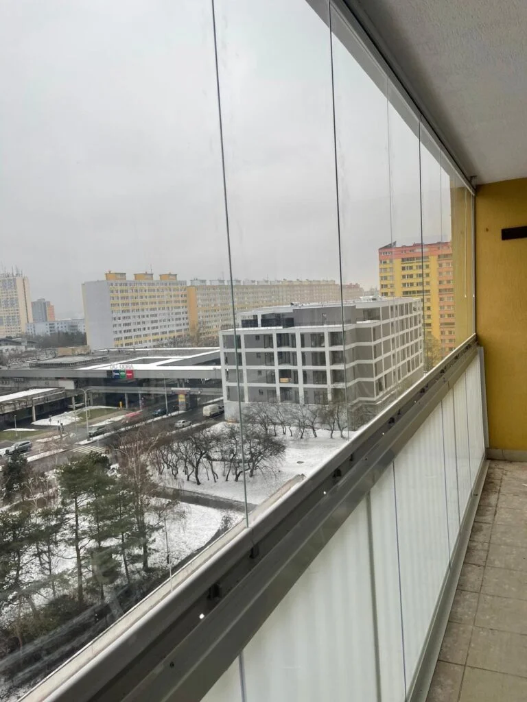 Zasklený balkon s výhledem na město v zimě.