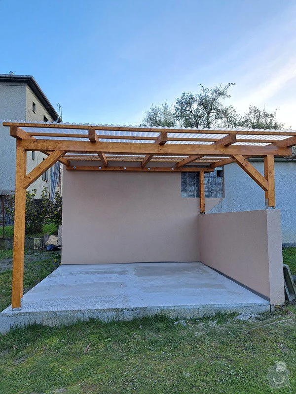 Nová dřevěná pergola s betonovou podlahou a dvěma stěnami v zahradním prostředí.