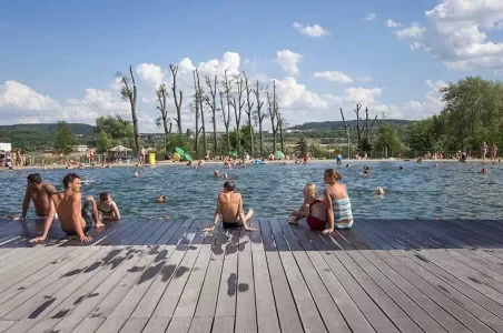 Lidé se koupají a odpočívají na dřevěné terase u bazénu.