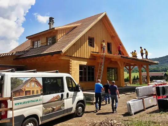Stavba roubené chalupy s dělníky na střeše pod modrou oblohou.