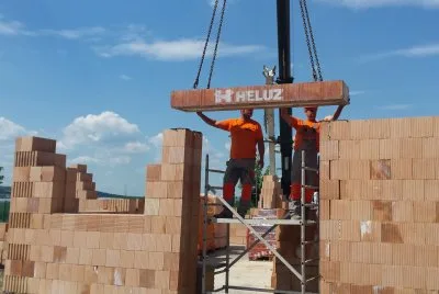 Výškový jeřáb zvedá cihly na staveništi s dělníky.