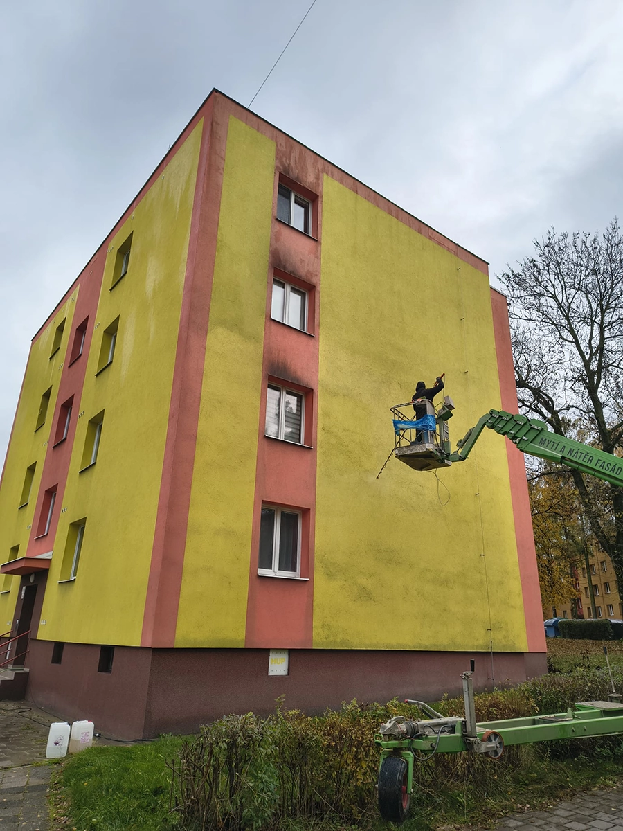 Pracovník maluje fasádu bytového domu žlutou barvou.